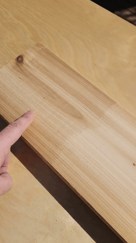 A piece of cedar with one half sanded and the other half unsanded. A person is pointing to the unsanded. The unsanded is rough and the sanded is smooth and ready for wood finish.