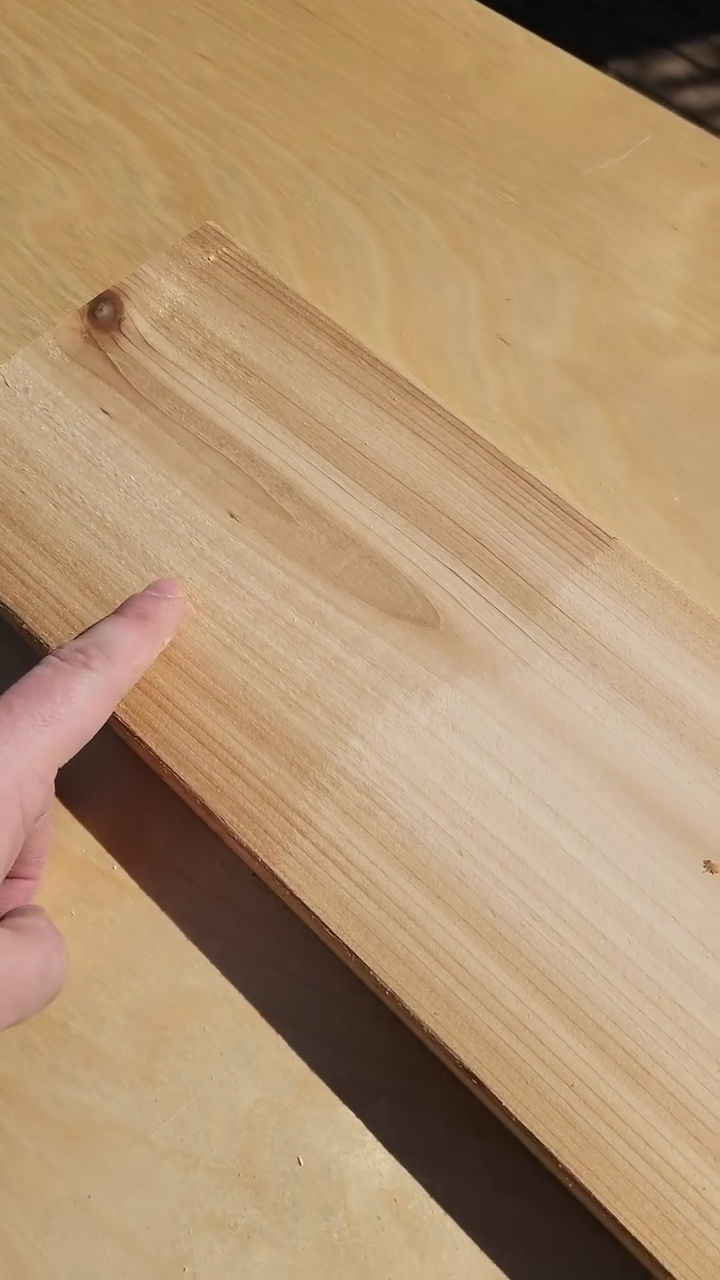 A piece of cedar with one half sanded and the other half unsanded. A person is pointing to the unsanded. The unsanded is rough and the sanded is smooth and ready for wood finish.